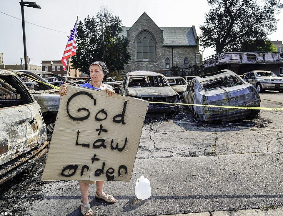 Жінка поряд зі згорілими авто тримає прапор США і табличку з написом: "Бог + закон + порядок"