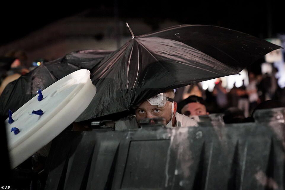 Протестующий прикрылся зонтиком и укрылся за мусорным контейнером во время столкновений у здания суда округа Кеноша