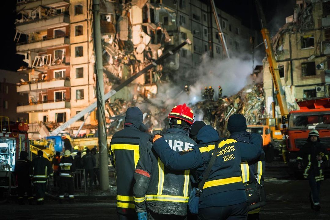 Спасатели и сотрудница психологической помощи обнимаются, глядя на уничтоженный Россией дом